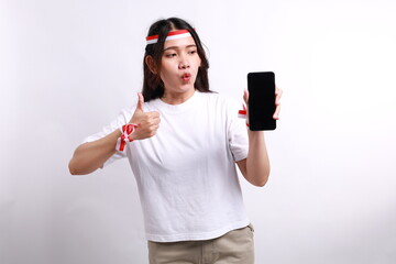 Surprised young Asian woman in casual t-shirt showing mobile phone with blank screen, showing thumb up gesture isolated on white background. indonesian independence day celebration concept