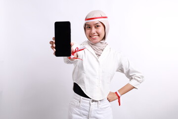 Excited young Asian Muslim woman showing a mobile phone with blank screen and celebrating success isolated over white background. Celebrate Indonesian independence day on 17 August