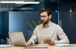 © Tetiana - A serious young businessman is sitting in the office at the desk, holding a tablet in his hand and working on a laptop with concentration