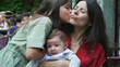 © Marco - Young girl giving a kiss to her mother while holding baby sibling. They share a joyful and loving family moment outdoors, displaying affection and happiness in a natural setting