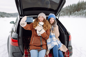 two women sitting in the trunk of her car and taking selfie on a background of snow-covered winter f