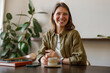 © Yaroslav Astakhov - A joyful woman sits at a table with coffee, surrounded by plants and books, exuding a warm atmosphere.