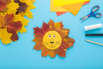 Wall Mural - Beautiful smiling lion head shape created from colorful maple leaves and paper on light blue table background. Pastel color. Making autumn decorations. Closeup. Top down view.