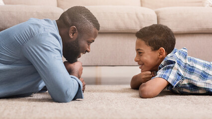 Wall Mural - Profile portrait of handsome african american man and his teen son lying on carpet at home and looking at each other