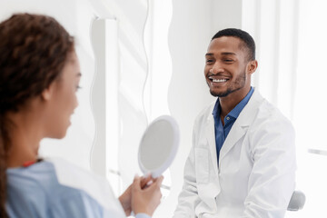 Black lady checking her white smile in mirror after stomatological treatment in modern clinic, happy dentist watching patient