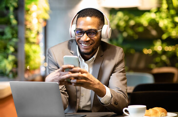 Enjoying Music. Happy afro businessman in suit wearing headphone and using his mobile phone sitting at cafe