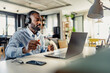 © lordn - Young African-American man wearing headphones, holding a coffee cup, and having a video call on his laptop in a modern home office setting.