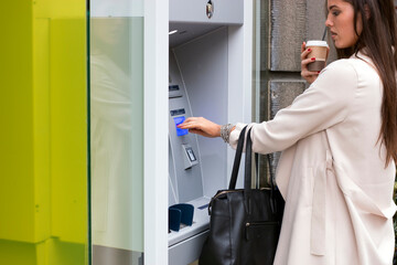 Attractive long haired brunette with shopping bags using automated teller machine while refreshing with coffee