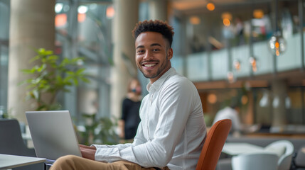 A young male employee is sitting on an office chair, smiling and looking at the camera while working with his laptop in front of him.