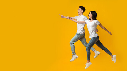 Excited asian couple pointing and running towards copy space, yellow background