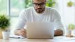 © NaphakStudio - focused man is working on laptop while taking notes, surrounded by greenery and office supplies. His concentration reflects productive work environment.