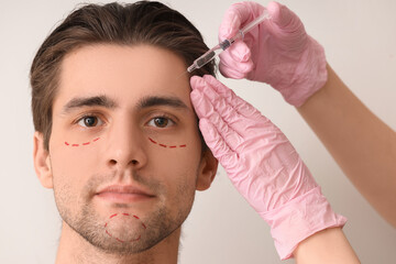 Young man with marked face receiving injection on light background, closeup. Plastic surgery concept