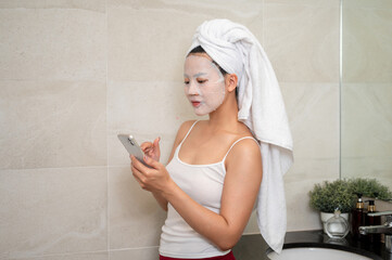 An Asian woman with a facial treatment mask on her face is using her smartphone in the bathroom.