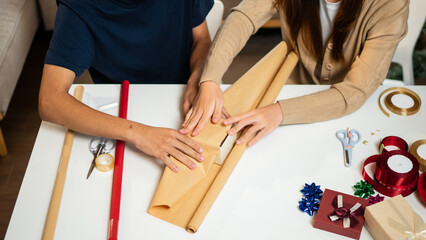 Wall Mural - A young man and woman are happily wrapping gifts at a table, sharing a fun moment together. They are surrounded by holiday decorations, with a Christmas tree adding to the festive ambiance