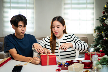 Wall Mural - A young man and woman are sitting at a table, happily wrapping gifts together. They share a joyful moment surrounded by holiday decorations.