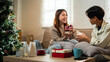 © snowing12 - A young couple sits together on a couch, exchanging gifts and sharing a warm holiday moment. The festive setting includes a Christmas tree and decorations, enhancing the cozy atmosphere