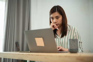 Asian woman is sitting at a desk with a laptop open in front of her. She is looking at the screen and she is deep in thought