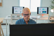 © Tj - Senior businessman wearing glasses focusing on computer screen with charts displayed on other monitors in bright modern office, indicating dedicated work and data analysis