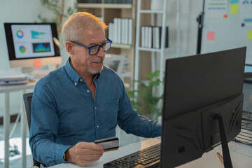 Senior businessman smiling while making an online payment with a credit card at his office desk, showcasing the seamless integration of business and technology