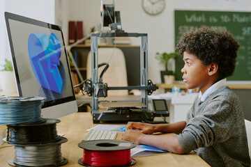 Young African American pupil inspecting design on computer in a modern classroom filled with technology and 3D printing equipment without looking at the camera