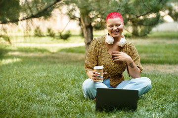 Wall Mural - A bald woman with bright red hair drinks coffee while smiling at her laptop outdoors in nature.