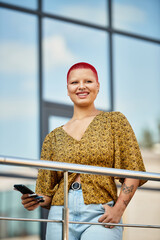 Wall Mural - A bald woman with striking red hair smiles while holding a phone on a sunny balcony.