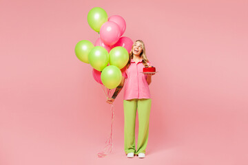 Full body young woman wears shirt celebrating birthday holiday and hold bunch of colorful air balloons, red cake with candles look aside on area isolated on plain pink background. Lifestyle concept.