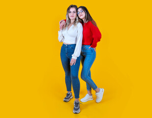 Two women together, full body view smiling happy friends posing over yellow studio background. Wearing jeans, sneakers and shirts. Blonde and brunette in fashionable hugging trendy 20s girls.