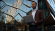 © Anastasia - A businessman in his 30s stands outside a modern building, holding a laptop and looking confident amid vibrant reflections and geometric architecture
