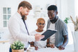 © BullRun - Confident estate agent showing documents to African American serious couple while standing in kitchen
