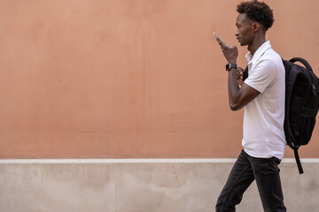 Young student walking and sending an audio message with his phone in front of a wall