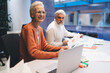© BullRun - Smiling senior businesswoman reviews documents in a modern office, sitting next to an attentive male colleague. Together, they work on project planning and data analysis, fostering productive team