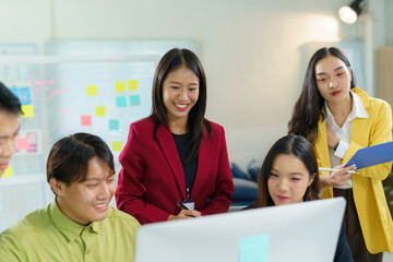 Group of young asian businesspeople discussing work using desktop computer in modern office meeting room with sticky notes on glass wall