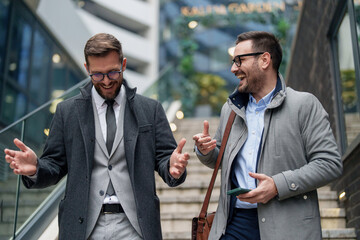 Two mid adult Caucasian businessmen engage in a lively discussion outdoors, dressed in smart casual attire, with one holding a smartphone.