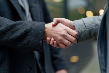 A close-up of a firm handshake between two mid-adult Caucasian men in business attire, signaling a professional agreement or greeting, set against an urban outdoor backdrop.