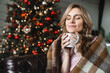 © InsideCreativeHouse - Caucasian woman wrapped warm blanket relaxing at home, sitting on cozy couch, happy smiling female drinking tea or coffee, dreaming, enjoying winter holidays with Christmas tree in the background