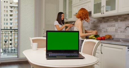 Laptop with a green screen on the table in the kitchen and two women cooking in the background.