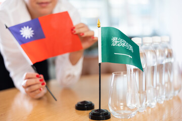 Unrecognizable woman preparing room for international negotiations and communication discussions of leaders. Lady sets miniatures flags of Saudi Arabia and Taiwan on table. Unfocused shot