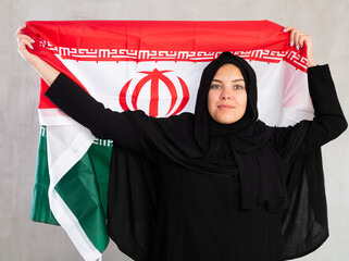 calm smiling Muslim woman in traditional black hijab holds flag of Iran. Portrait of female Muslim with Iranian flag on gray background