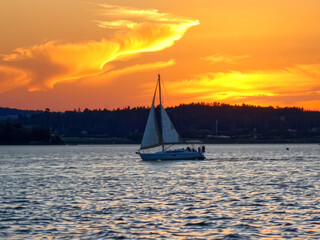 A sailboat is sailing on a calm lake at sunset. The sky is filled with clouds and the sun is setting, creating a beautiful and serene atmosphere