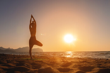 Vrikshasana tree pose from yoga, female silhouette at sunset or sunrise. Back view of a girl practicing morning workout on the seashore