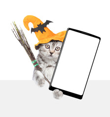 Poster - Cute kitten wearing hat for halloween holds shows smartphone with white blank screen in it paw above white blank board. Empty space for text. Isolated on white background