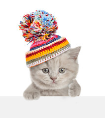 Poster - Cute kitten wearing a warm hat with pompon looks above blank banner and shows thumbs up gesture. isolated on white background