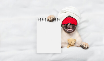 Fototapete - Pug puppy wearing sleeping mask lying with favorite toy bear under white blanket on a bed at home and showing blank notepad. Top down view. Empty space for text
