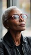 © Denisa - An older Black woman in transparent frames looks thoughtfully by a window in soft light