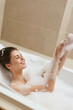 © SHOTPRIME STUDIO - Joyful young woman enjoying a bubble bath, laughing and playing with suds, creating a relaxed and cheerful atmosphere in a serene bathroom setting.