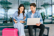 © Iryna - Caucasian male and female young adults sitting on bench with laptop and pink suitcase working together, sharing ideas in modern outdoor setting.