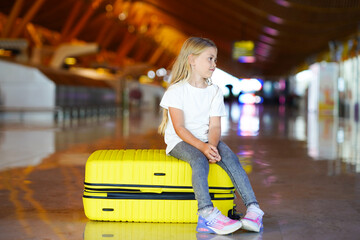 A young girl sitting on a bright yellow suitcase, waiting in a spacious airport terminal with modern architecture.
