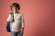© Guys Who Shoot - in a photo studio on a pink background stands a smiling boy of ten years old, a schoolboy in a white sweater with a blue briefcase preparing for school little tourist