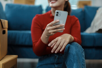 Young woman using smartphone while sitting on floor near sofa and cardboard boxes, relaxing after moving into new home